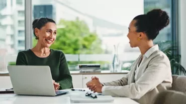 Deux femmes échangent autour d’une table, un ordinateur portable ouvert entre elles.