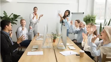 Mensen applaudisseren en lachen in een vergaderruimte rond een tafel.