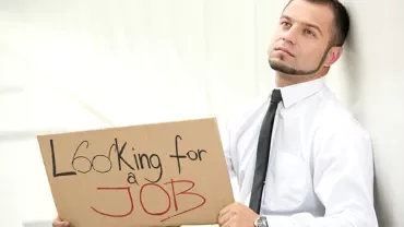 Person holding a cardboard sign reading 'Looking for a Job' while sitting against a wall.