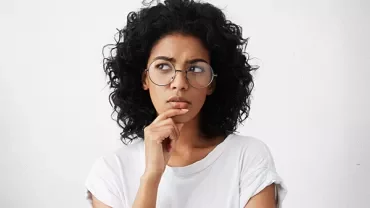 Personne aux cheveux bouclés portant des lunettes, d'apparence pensive.