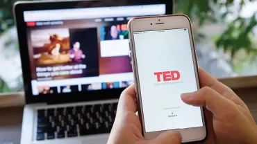 Persoon gebruikt TED-app op smartphone met laptop op de achtergrond.