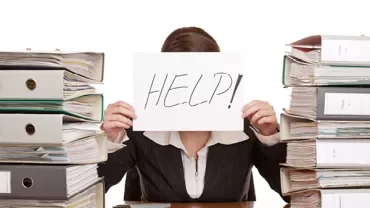 Person behind desk with 'HELP!' sign, surrounded by files.