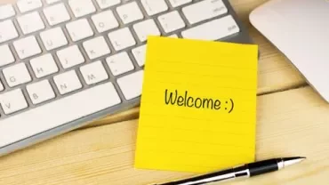 Note jaune avec 'Welcome :)' sur un bureau en bois à côté d'un clavier et d'un stylo.