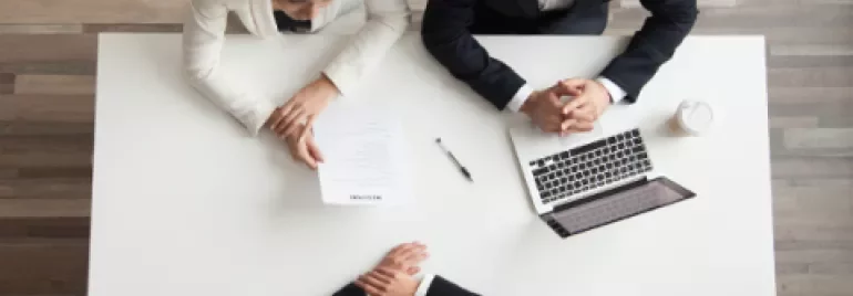 Drie mensen aan tafel tijdens een vergadering met een laptop en documenten.