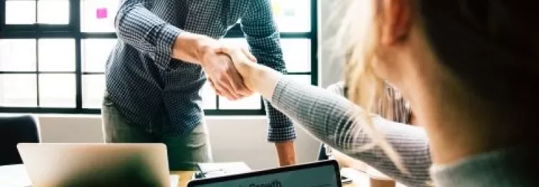 People shaking hands in an office setting with a laptop open on the table.