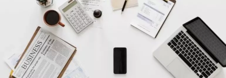 Kantoorruimte met laptop, krant, koffie en rekenmachine op een bureau.