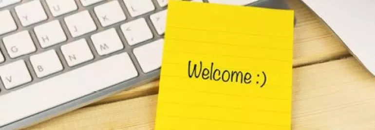 Yellow sticky note with 'Welcome :)' on a wooden desk beside a keyboard and pen.