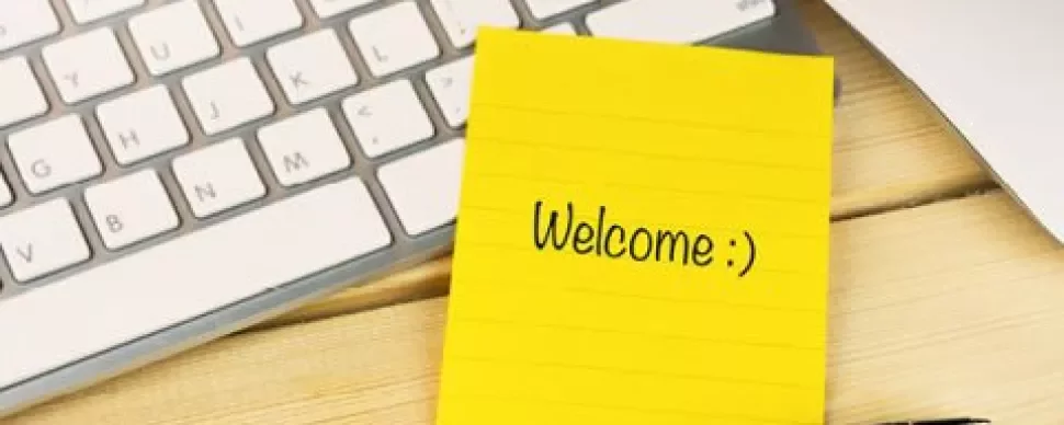 Note jaune avec 'Welcome :)' sur un bureau en bois à côté d'un clavier et d'un stylo.