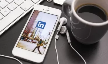 Smartphone met LinkedIn-app, oordopjes, koffiemok op bureau.